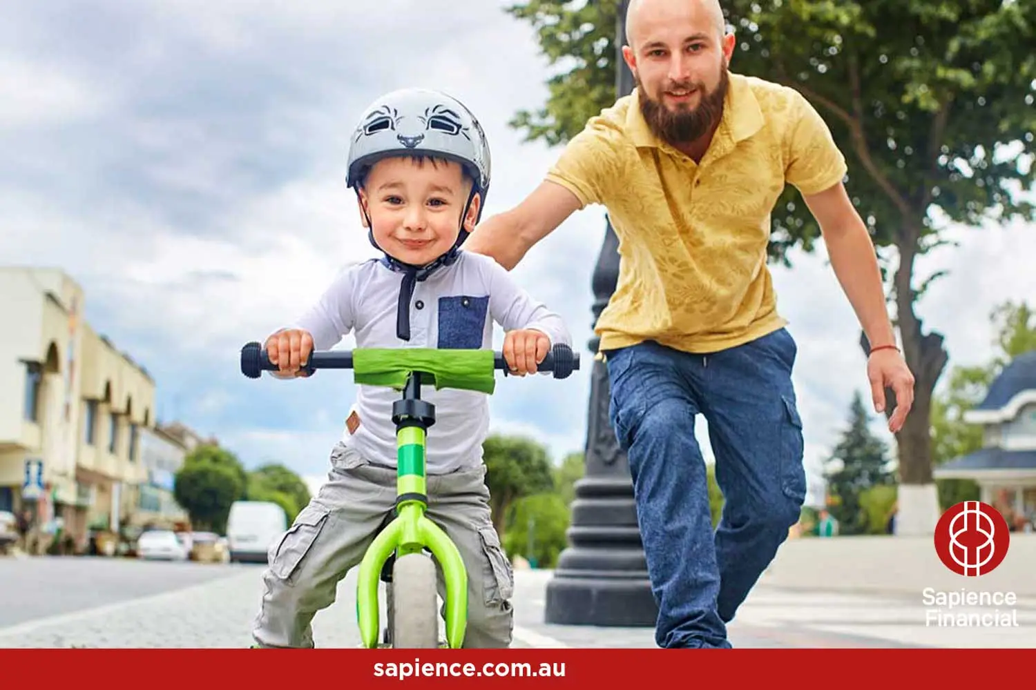 father in yellow shirt pushing his young son on a green bicycle wearing a safety helmet as he learns how to ride a bike