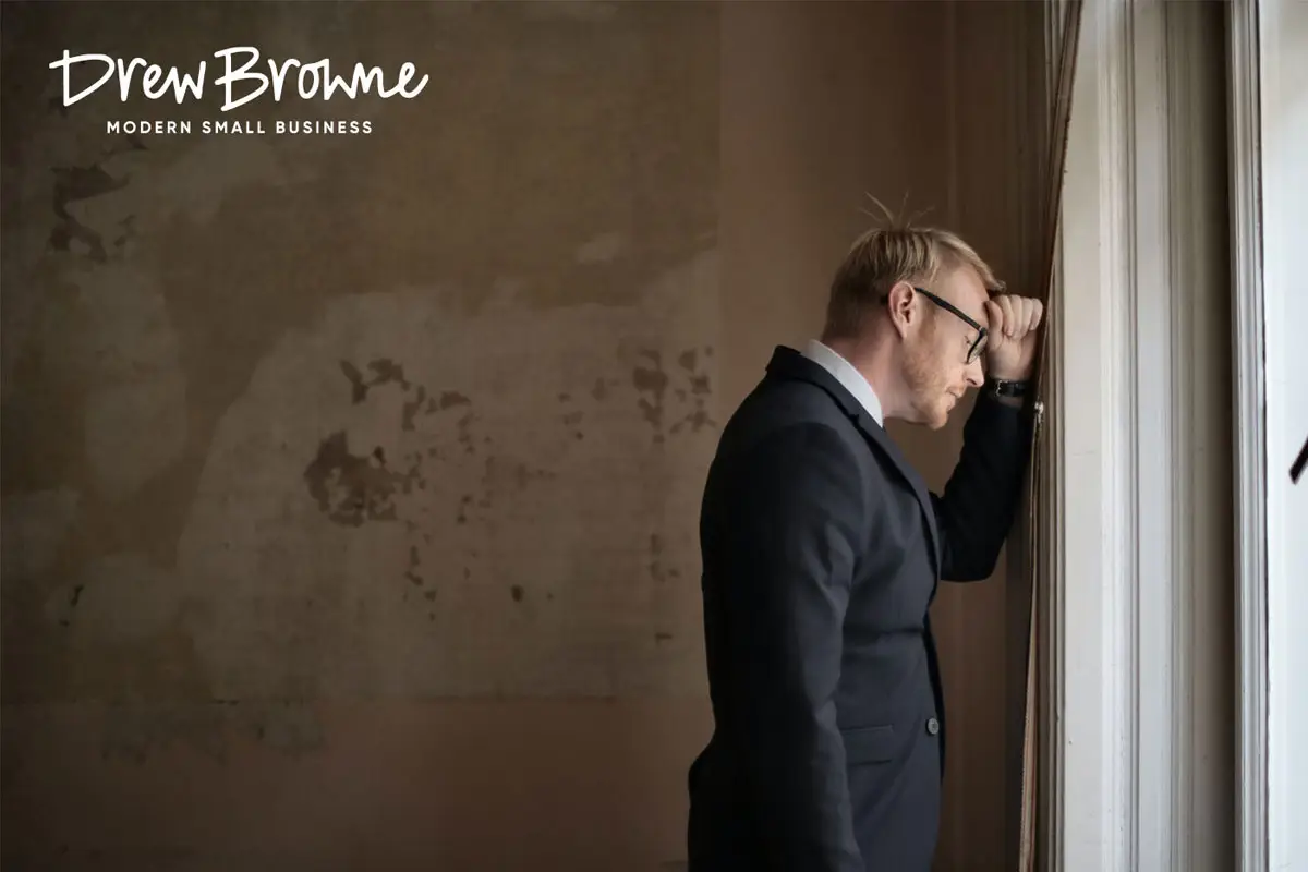 A middle aged businessman with glasses looking out of a window with his full head on his arm in deep thought